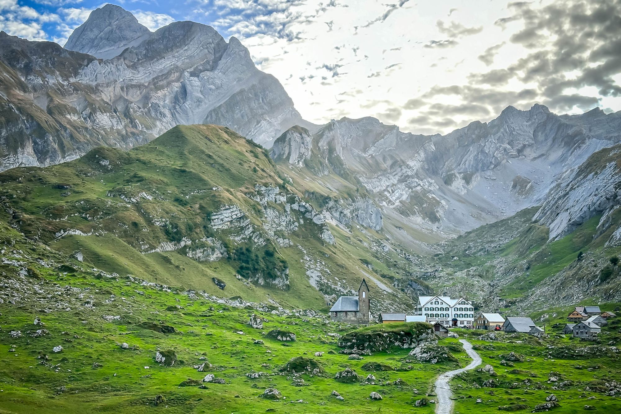 A footpath leading through green alpine meadows to a small mountain settlement beneath limestone peaks.